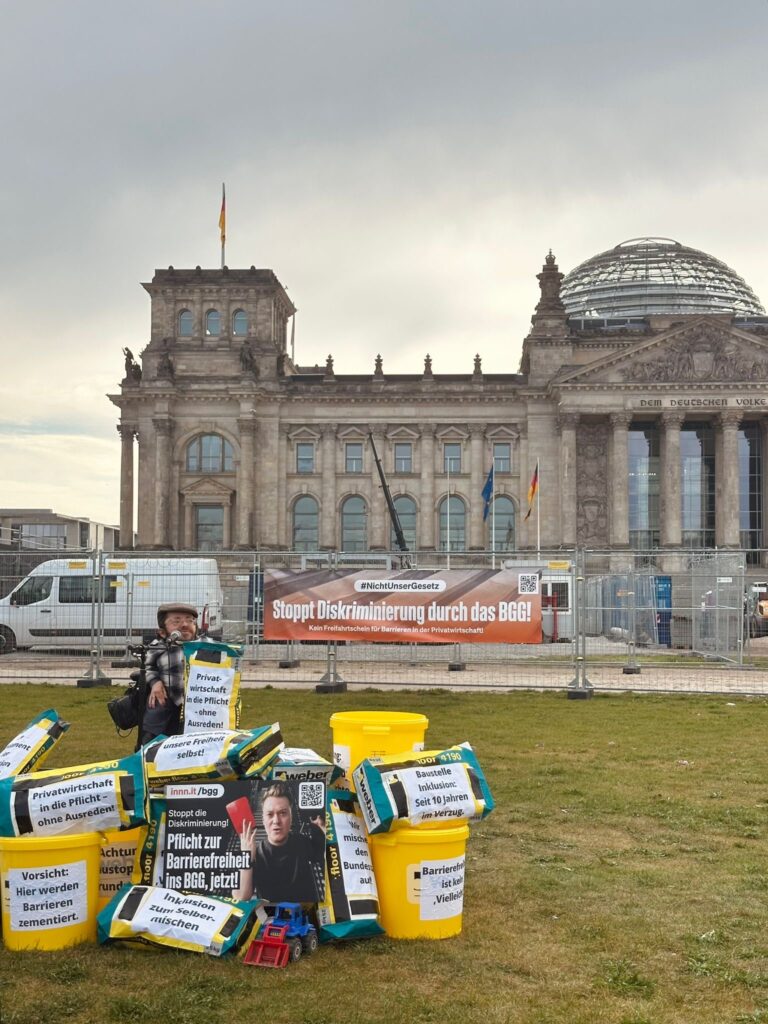Raul Krauthausen spricht vor dem Bundestag bei einer Demo, Vor ihm sind Eimer, und Zementsäcke gestapelt.