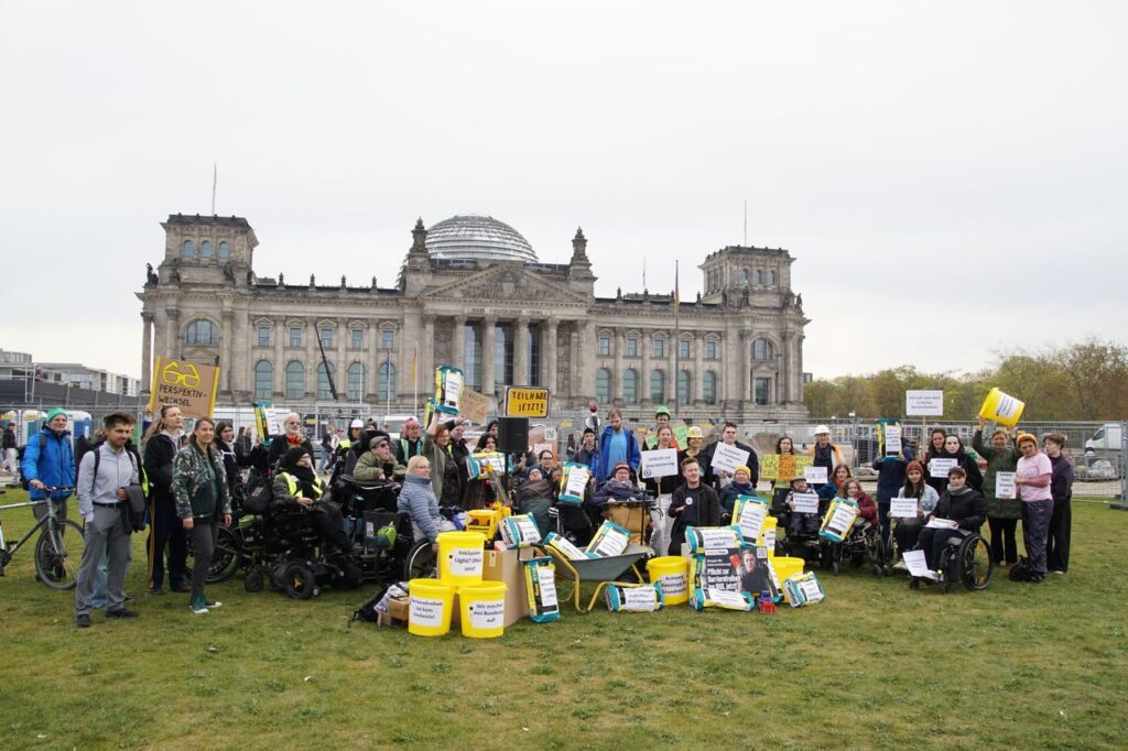 Demo vor dem Bundestag, Gruppenfoto der Demonstrierenden mit und ohne Rollstuhl, zu sehen sind Eimer, Schubkarren, Zementsäcke
