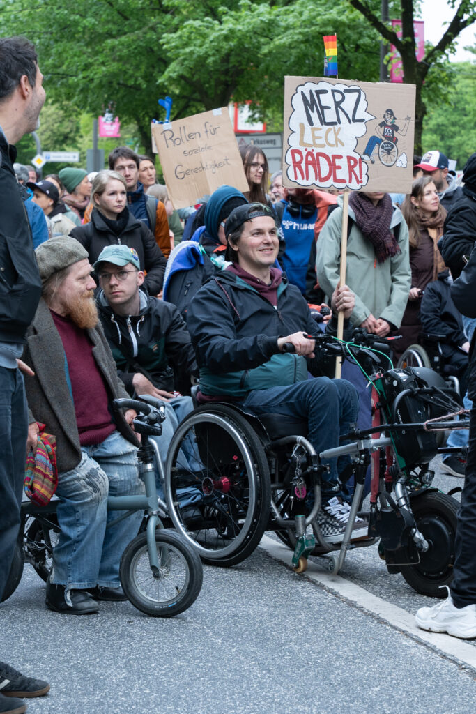 Mehrere Teilnehmende stehen und sitzen während der Demonstration zusammen. Im Mittelpunkt hält ein Rollstuhlfahrer ein Schild mit der Aufschrift: „Merz leck Räder!“ Dahinter ist ein weiteres Schild zu sehen: „Rollen für soziale Gerechtigkeit“.