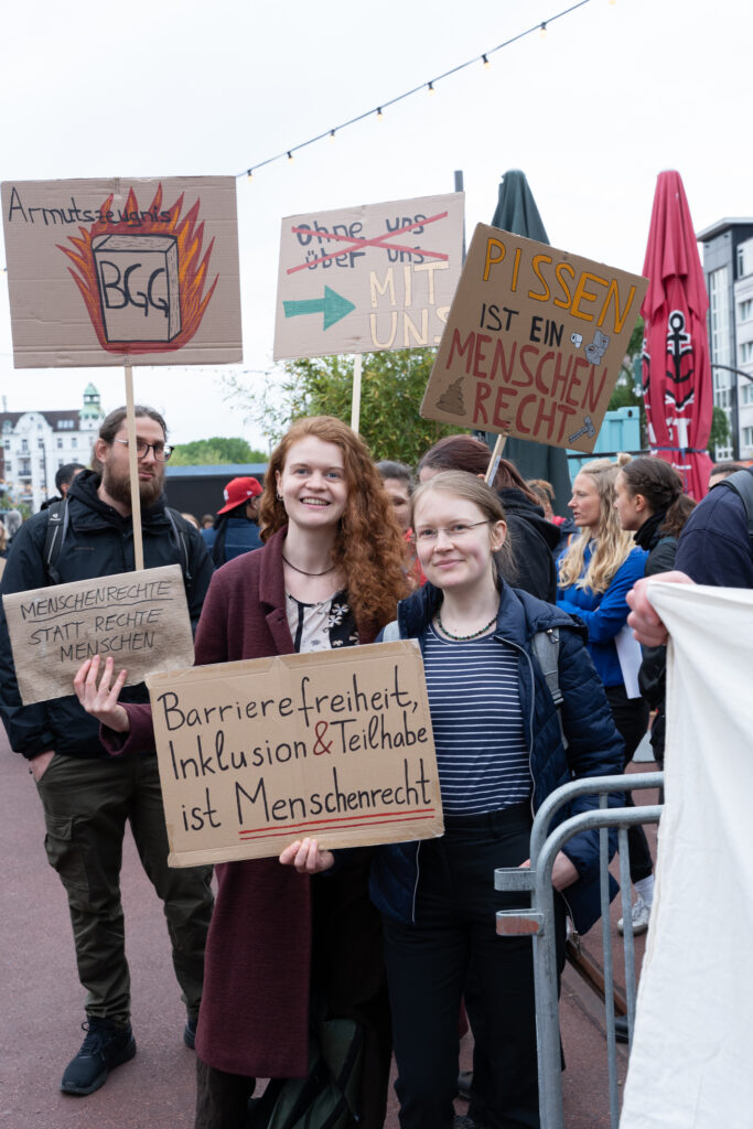 Drei Menschen posieren mit selbstgemalten Protestschildern. Auf den Schildern stehen unter anderem: „Barrierefreiheit, Inklusion & Teilhabe ist Menschenrecht“, „Menschenrechte statt rechte Menschen“, „Pissen ist ein Menschenrecht“ und „ohne über uns – mit uns“.