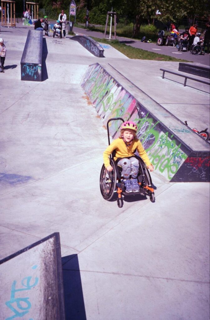 Ein Kind mit pinkem Helm fährt im Rollstuhl durch den Skatepark. Im Hintergrund warten und skaten weitere Kinder und Erwachsene, einige davon ebenfalls im Rollstuhl.