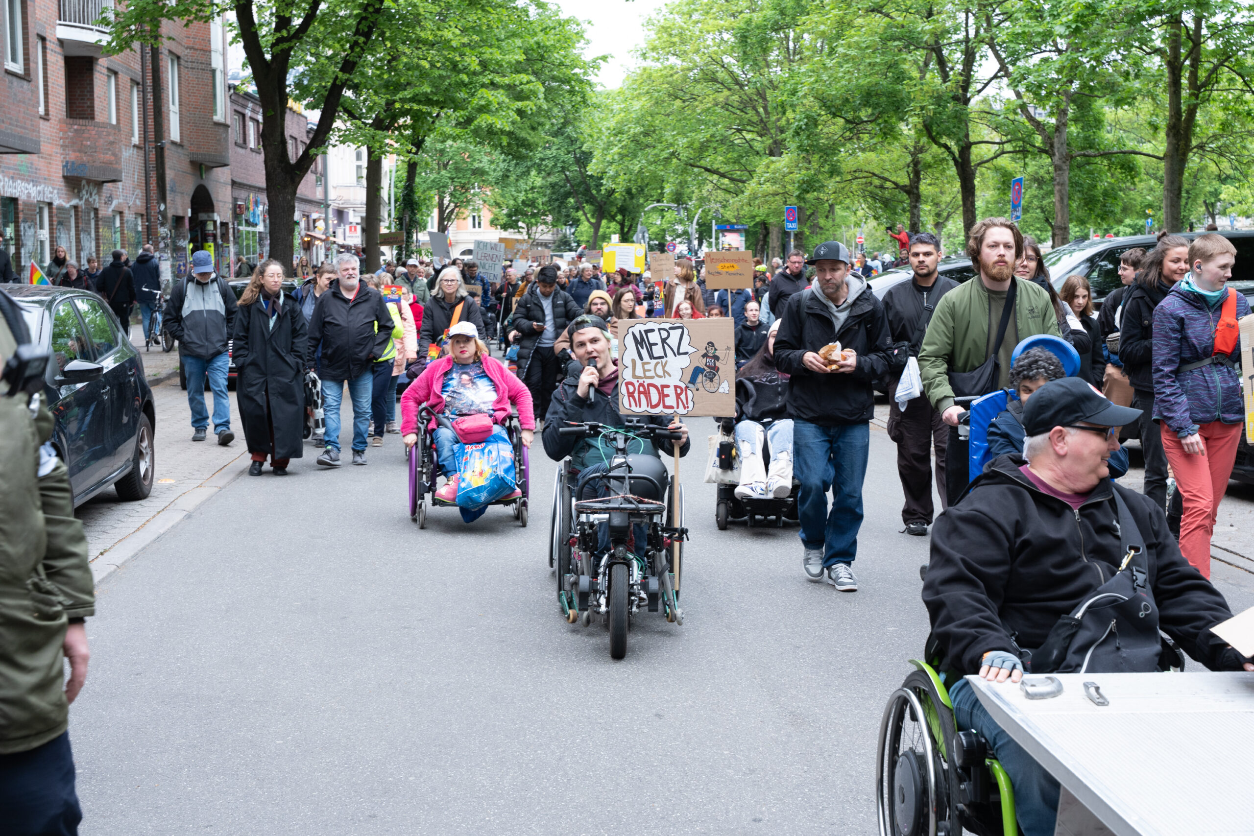 Ein großer Demonstrationszug zieht durch eine enge, grüne Straße auf St. Pauli. Im Vordergrund sind mehrere Rollstuhlfahrende und viele weitere Teilnehmende mit Protestschildern zu sehen. Ein Schild trägt die Aufschrift: „Merz leck Räder!“