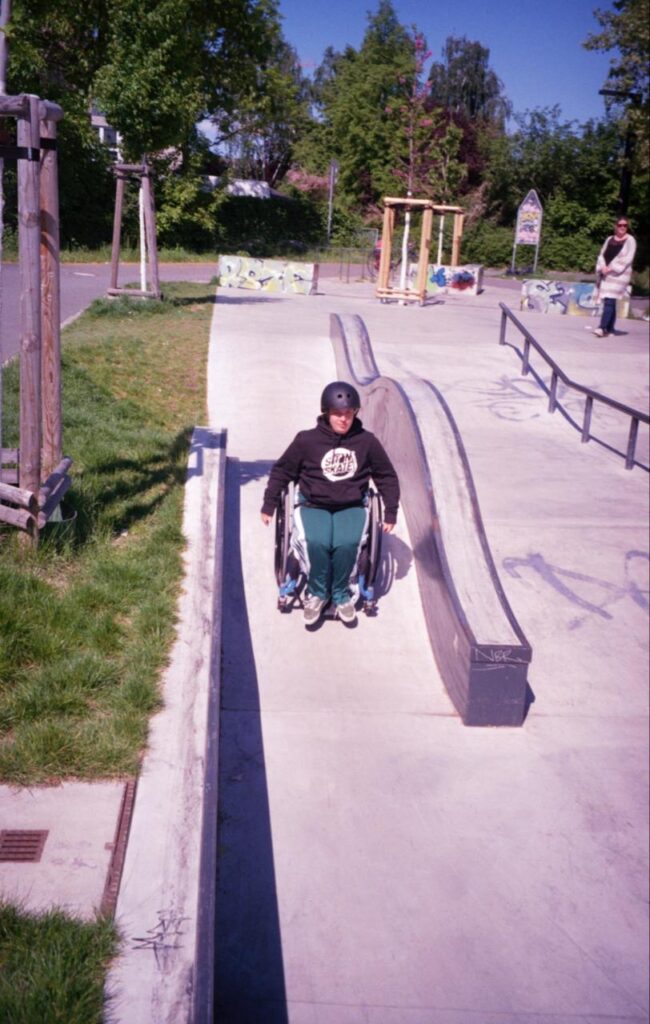 Eine Rollstuhlfahrende Person mit Helm fährt durch einen schmalen, abgesenkten Bereich neben einer langen Beton-Ledge. Der sonnige Skatepark ist von Bäumen und Grünflächen umgeben.