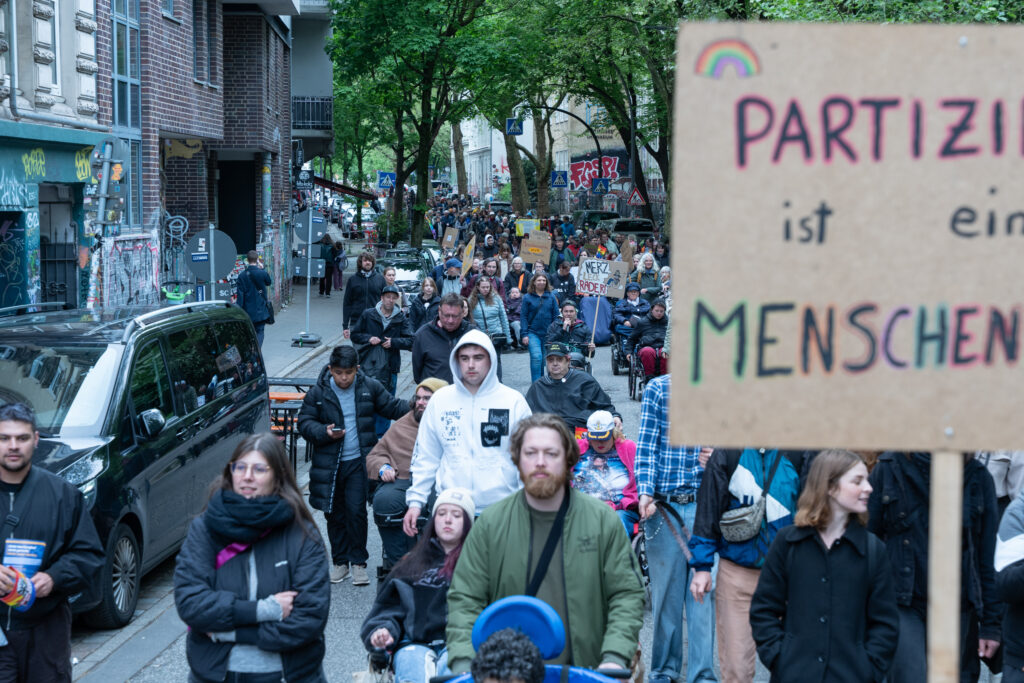 Blick auf einen langen Demonstrationszug in einer engen Straße auf St. Pauli. Die Menschen laufen dicht hintereinander, viele mit Schildern. Im Vordergrund ist unscharf ein großes Schild zu sehen, im Hintergrund wird die Länge des Demozugs sichtbar.