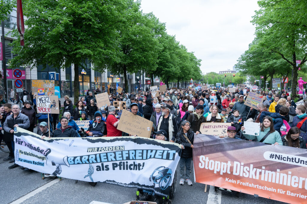 Große Menschenmenge auf der Reeperbahn hinter zwei Bannern. Auf einem Banner steht: „Wir fordern Barrierefreiheit als Pflicht!“ Auf dem anderen steht: „Stoppt Diskriminierung“. Viele Teilnehmende halten Schilder hoch.