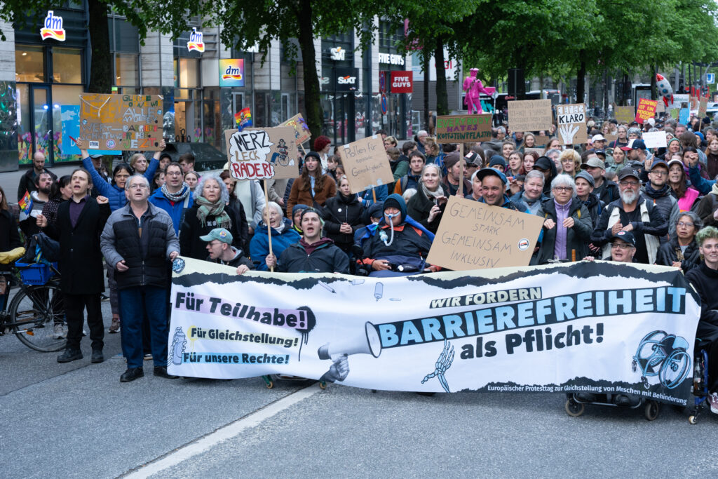 Nahaufnahme der Demonstration auf der Reeperbahn. Viele Menschen mit und ohne Behinderung stehen dicht zusammen hinter einem großen Banner mit der Aufschrift: „Für Teilhabe! Für Gleichstellung! Für unsere Rechte! Wir fordern Barrierefreiheit als Pflicht!“ Einige rufen Parolen und halten Protestschilder hoch.