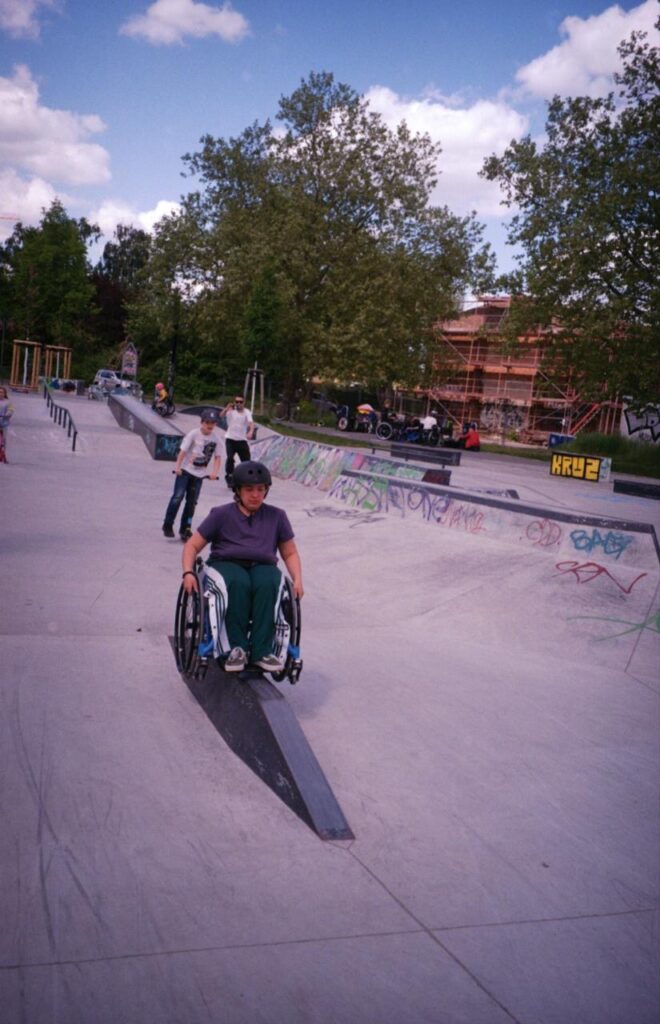 Eine Rollstuhlfahrende Person fährt über ein niedriges, keilförmiges Skateelement. Im Hintergrund sind weitere Skatende und mehrere Rollstuhlfahrende im Skatepark zu sehen.