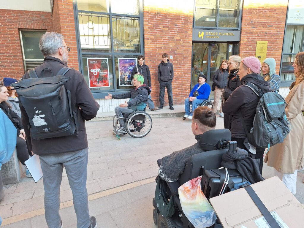 Eine Gruppe steht vor einem Gebäude in Ottensen und hört einer Person im manuellen Rollstuhl zu, die eine Barriere im Alltag erklärt. Im Vordergrund sitzt eine weitere Person in einem E-Rollstuhl. Die Teilnehmenden sind im Gespräch über Zugänglichkeit und Barrieren im Stadtteil.