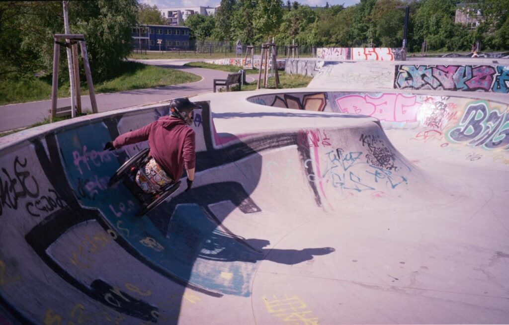Ein Rollstuhlskater fährt hoch an der gebogenen Wand einer Bowl. Der betonierte Skatepark ist mit Graffiti bedeckt.
