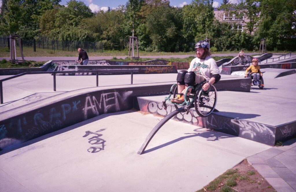 Ein Rollstuhlskater mit Helm und Schonern fährt über ein gebogenes Metallelement im Skatepark. Im Hintergrund skatet ein Kind im Rollstuhl zwischen Rampen und Rails.