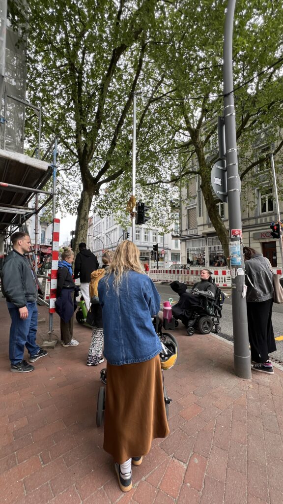 Eine Gruppe von Menschen bei einem Barrierespaziergang an einer Straßenkreuzung in Ottensen. Eine Person im E-Rollstuhl spricht mit der Gruppe. Baustellenabsperrungen, Gerüste und ein schmaler Gehwegbereich zeigen die Herausforderungen im öffentlichen Raum.