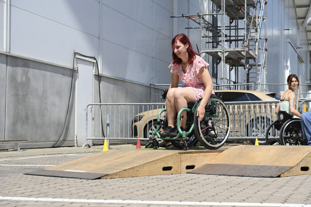 Eine rothaarige Rollstuhlfahrerin fährt lächelnd über eine Rampe des mobilen SIT’N’SKATE-Skateparks vor der Hamburger Messehalle.