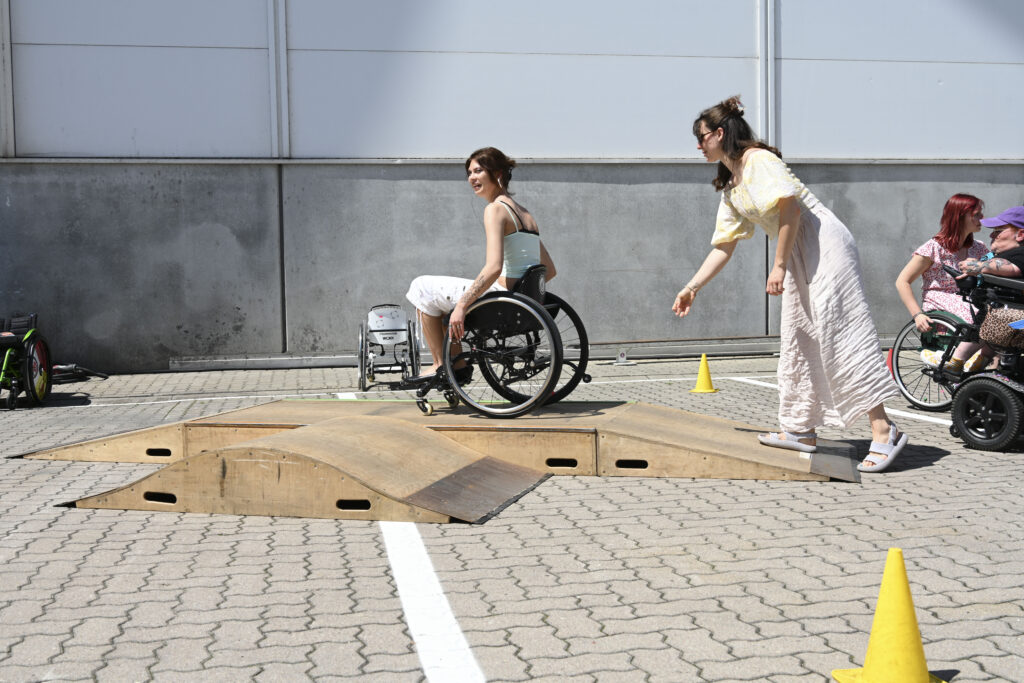 Eine Rollstuhlfahrerin fährt über den mobilen Skatepark, während eine Person neben der Rampe begleitet. Im Hintergrund sind weitere Messebesucher*innen im Rollstuhl zu sehen.