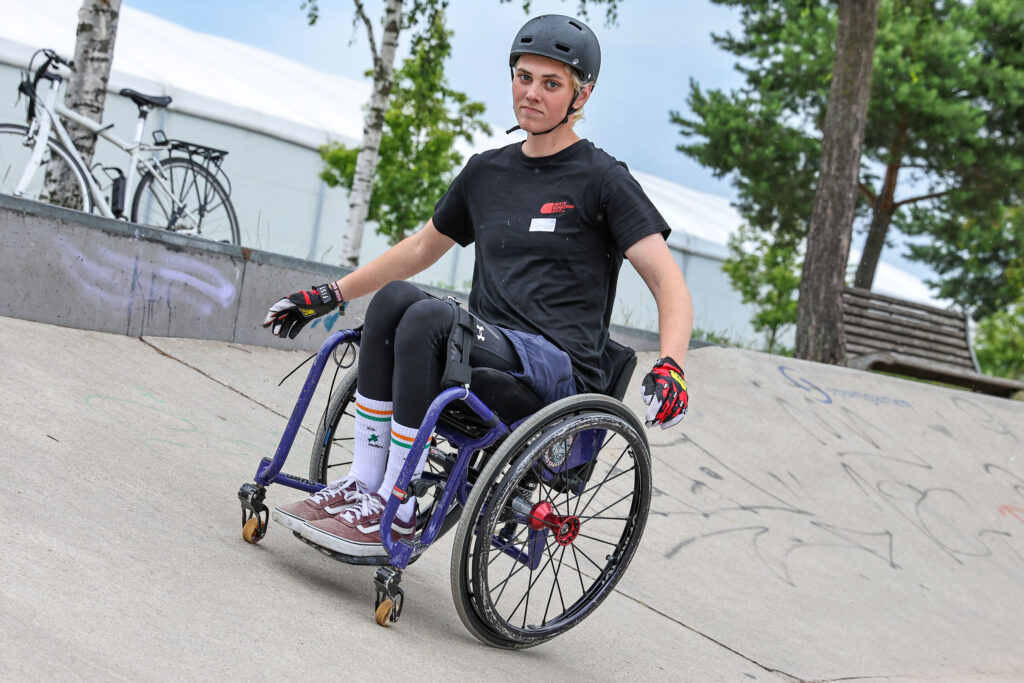 Eine Person mit Helm und Handschuhen fährt im lila Rollstuhl eine schräge Betonrampe im Skatepark hinunter. Im Hintergrund stehen Bäume, Fahrräder und ein großes weißes Zelt.