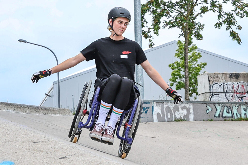 Eine Person mit schwarzem Helm fährt im lila Rollstuhl schräg über eine Betonrampe im Skatepark. Die Person hält die Arme seitlich ausgestreckt, im Hintergrund sind Graffiti, ein Baum und Gebäude zu sehen.