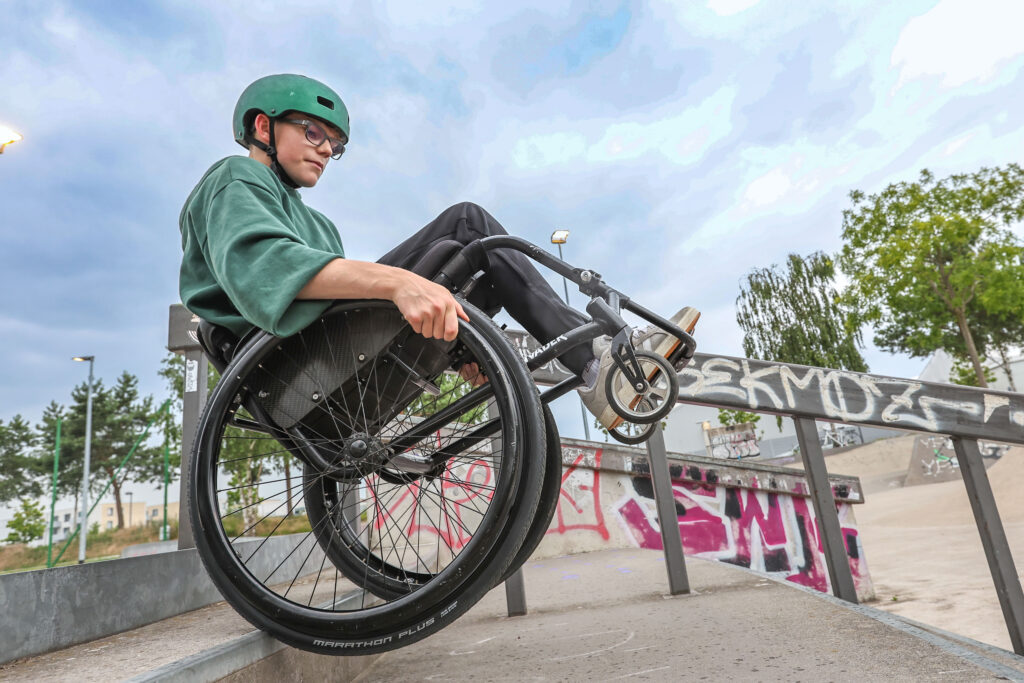 Eine Person mit grünem Helm und Brille balanciert mit dem Rollstuhl an einem Metallgeländer im Skatepark. Die Aufnahme zeigt die Aktion aus niedriger Perspektive vor einem wolkigen Himmel.