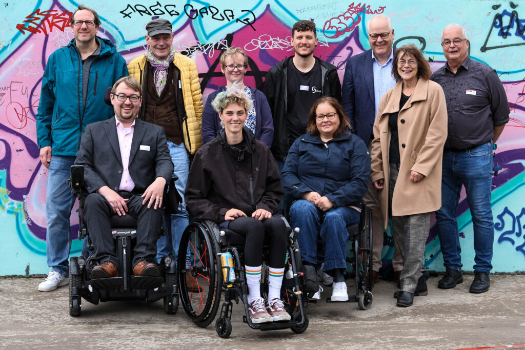 Gruppenfoto von elf Menschen vor einer bunt mit Graffiti besprühten Wand. Drei Personen sitzen vorne in Rollstühlen, die übrigen stehen dahinter.