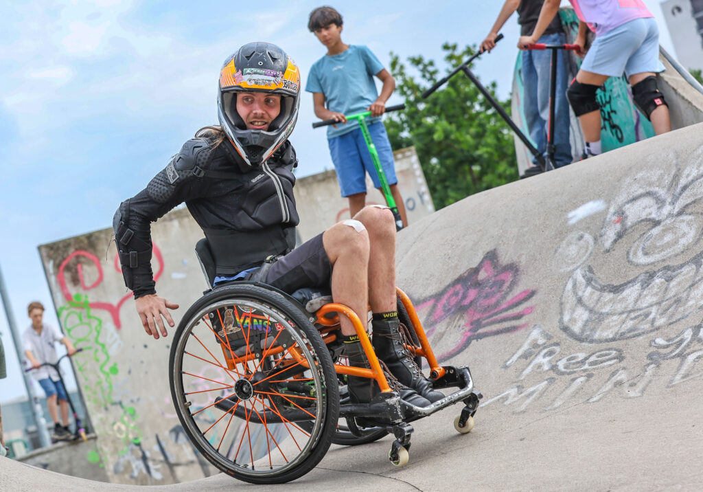 Ein Rollstuhlskater mit Integralhelm und Schutzkleidung fährt mit einem orangefarbenen Skate-Rollstuhl eine schräge Betonrampe hinunter. Im Hintergrund beobachten mehrere Jugendliche mit Scootern die Aktion.