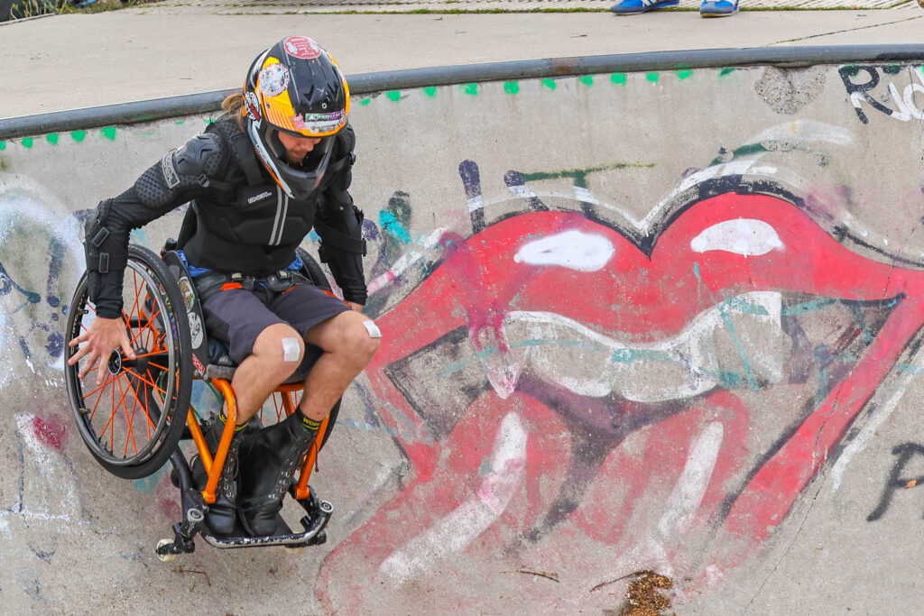 Ein Rollstuhlskater mit Integralhelm und Schutzkleidung fährt mit einem orangefarbenen Skate-Rollstuhl durch eine mit Graffiti bemalte Betonbowl.