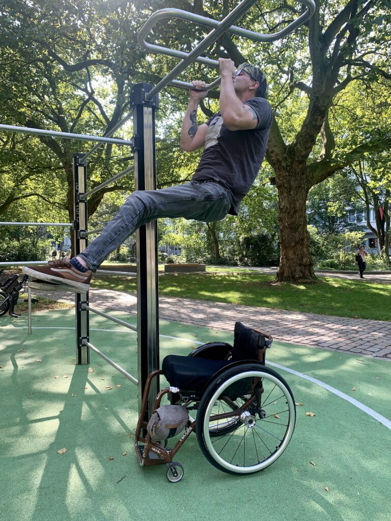 ein Mann macht einen Klimmzug, unter ihm steht sein Rollstuhl