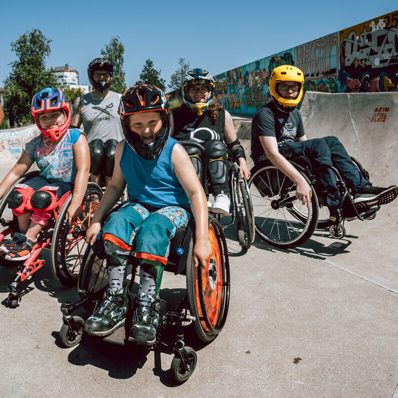 Eine Gruppe von Rollstuhlfahrern und Rollstuhlfahrerinnen mit Helmen im Skatepark, Kinder, Jugendliche und Erwachsene.