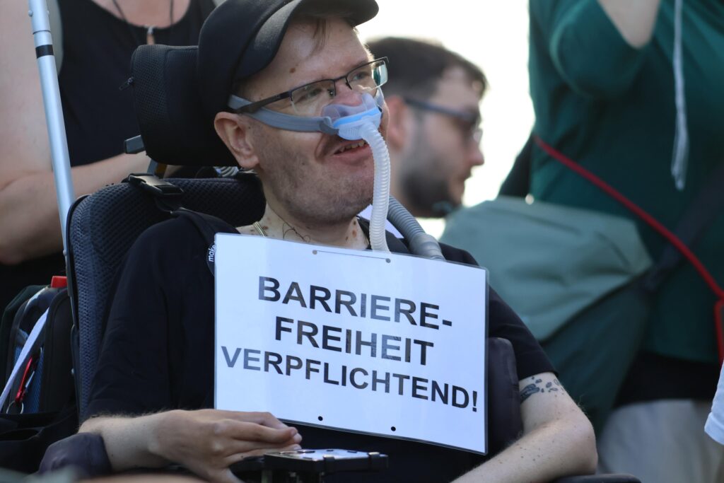 Eine Person im Elektrorollstuhl trägt eine Atemmaske und ein Schild mit der Aufschrift „Barrierefreiheit verpflichtend!“.