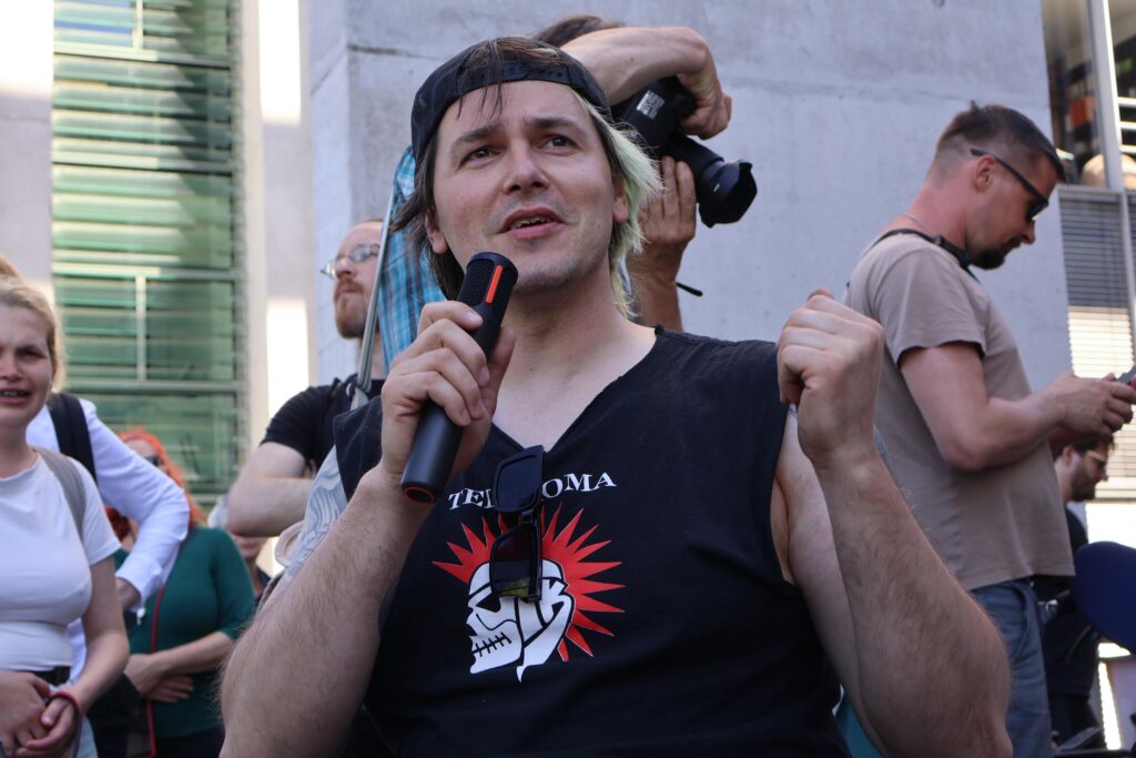 Ein Mann in schwarzem ärmellosem Shirt spricht bei der Demonstration in ein Mikrofon und gestikuliert mit der freien Hand. Hinter ihm stehen weitere Teilnehmende.