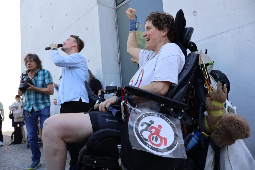 Eine Person im Elektrorollstuhl hebt bei der Demonstration lächelnd die Faust. Im Hintergrund spricht ein Mann in ein Mikrofon.