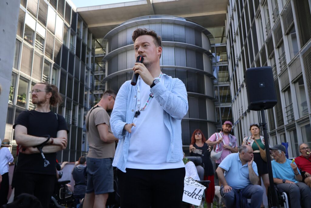 Ein junger Mann spricht mit einem Mikrofon zu den versammelten Demonstrierenden. Im Hintergrund sind Menschen im Rollstuhl und weitere Teilnehmende zu sehen.