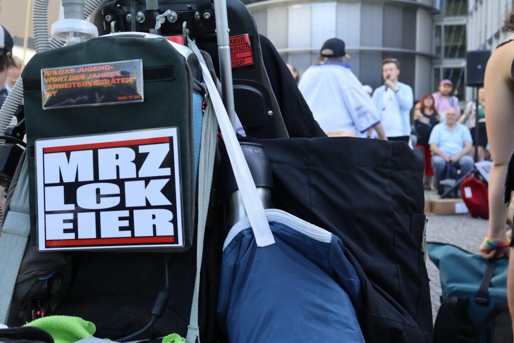 Nahaufnahme der Rückseite eines Elektrorollstuhls mit politischen Aufklebern. Einer trägt die große Aufschrift „MRZ LCK EIER“, darüber steht: „Bis das Jugendwort des Jahres Arbeiterverräter ist“.