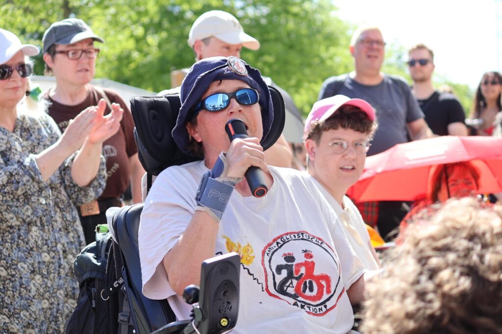 Eine Person im Elektrorollstuhl spricht bei der Demonstration in ein Mikrofon. Rundherum stehen weitere Menschen, einige applaudieren.