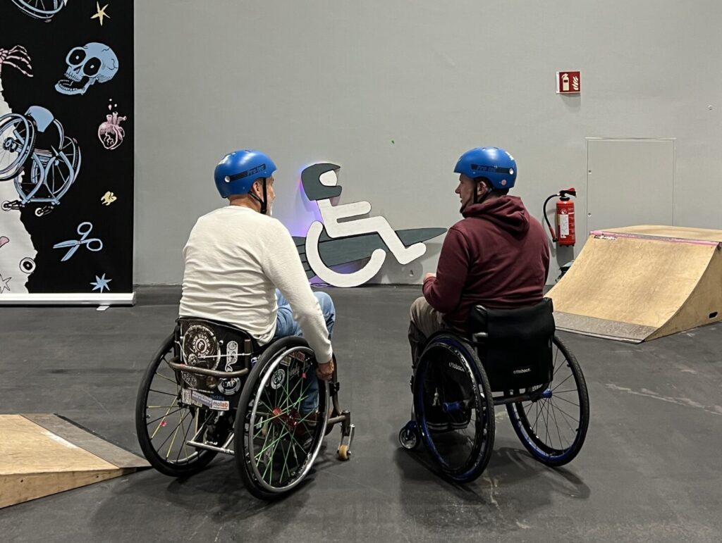Zwei Rollstuhlfahrer sitzen zwischen den Elementen des mobilen SIT’N’SKATE-Skateparks in einer Messehalle und sprechen miteinander. Im Hintergrund stehen eine kleine Rampe und eine große Rollstuhl-Grafik.