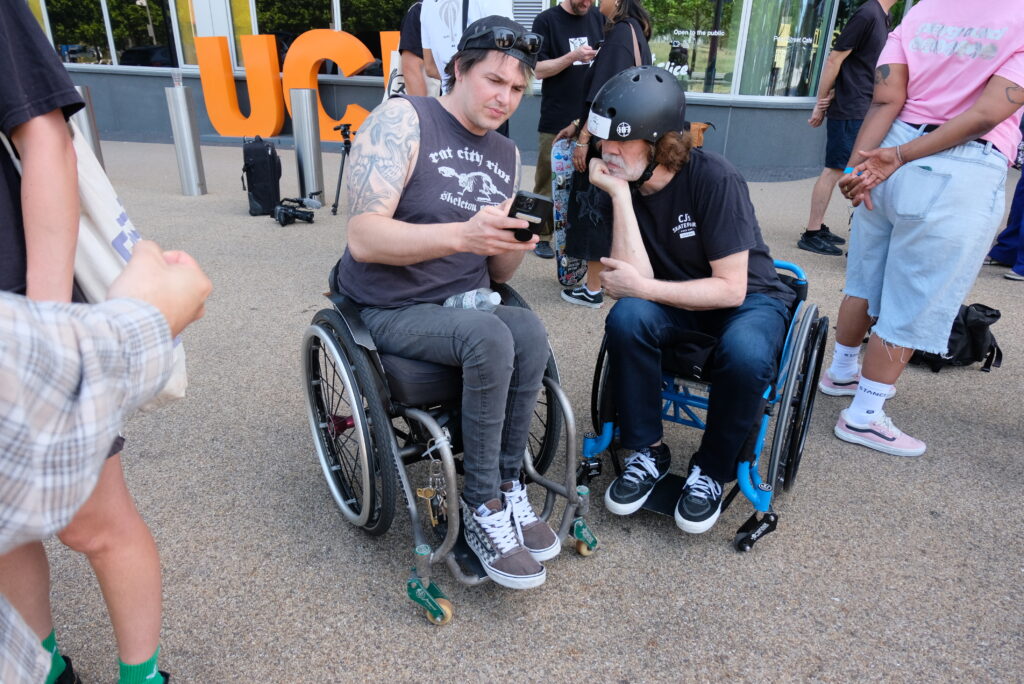 David zeigt einem anderen Rollstuhlfahrer auf dem Vorplatz der UCL etwas auf seinem Smartphone. Beide sitzen im Rollstuhl und sind von weiteren Teilnehmenden des Social Skate Summit umgeben. Im Hintergrund sind das Universitätsgebäude und große orangefarbene Buchstaben der UCL zu sehen.