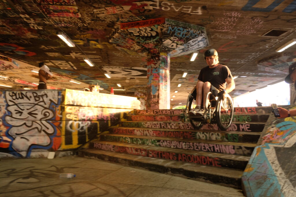 Ein Rollstuhlskater fährt in einem stark mit Graffiti gestalteten Skatepark eine Treppe hinunter. Die Szene wird von warmem Abendlicht beleuchtet. Im Hintergrund und am Rand des Skateparks beobachten andere Personen die Aktion.