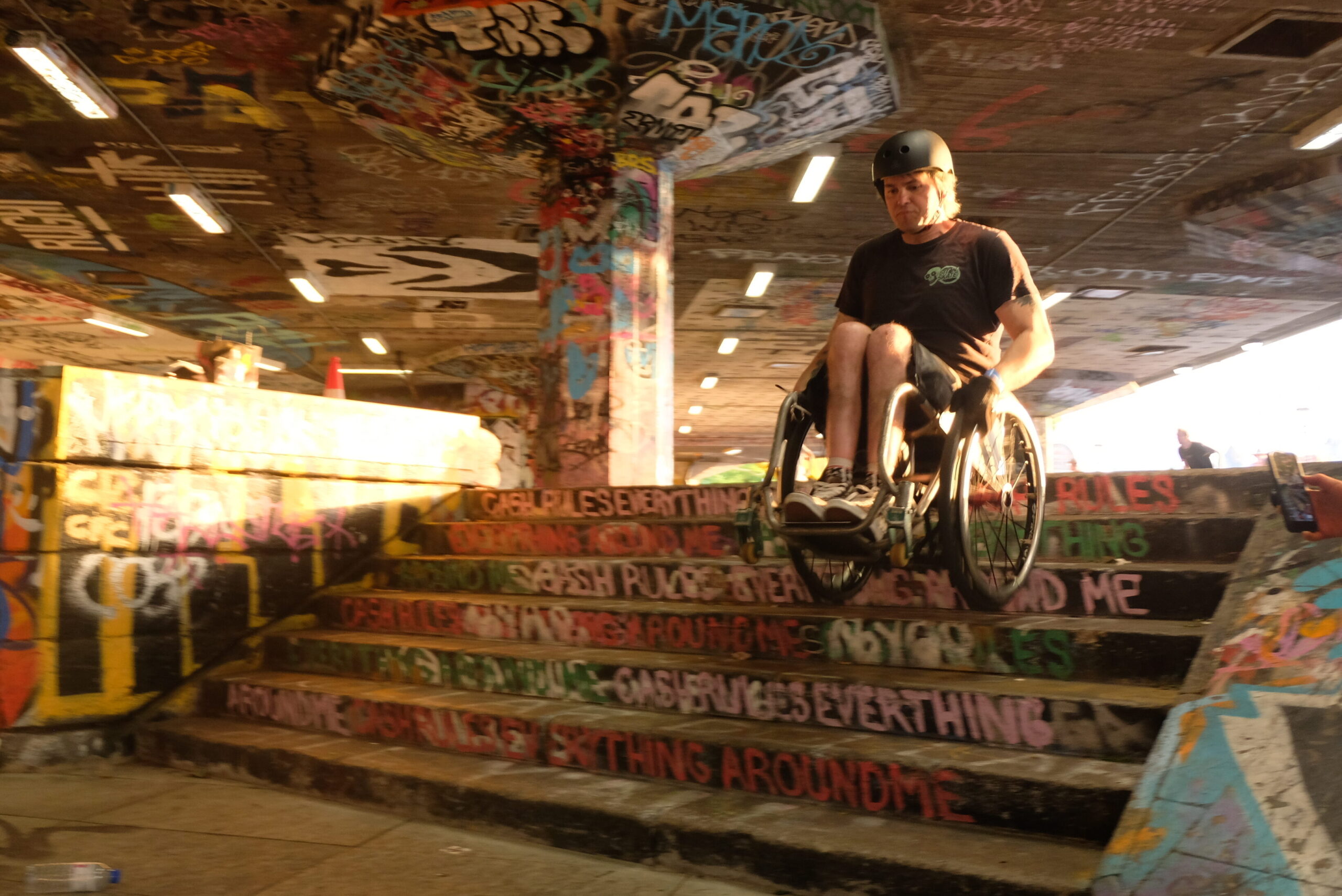 Ein Rollstuhlskater fährt in einem stark mit Graffiti gestalteten Skatepark eine Treppe hinunter. Die Szene wird von warmem Abendlicht beleuchtet. Im Hintergrund und am Rand des Skateparks beobachten andere Personen die Aktion.