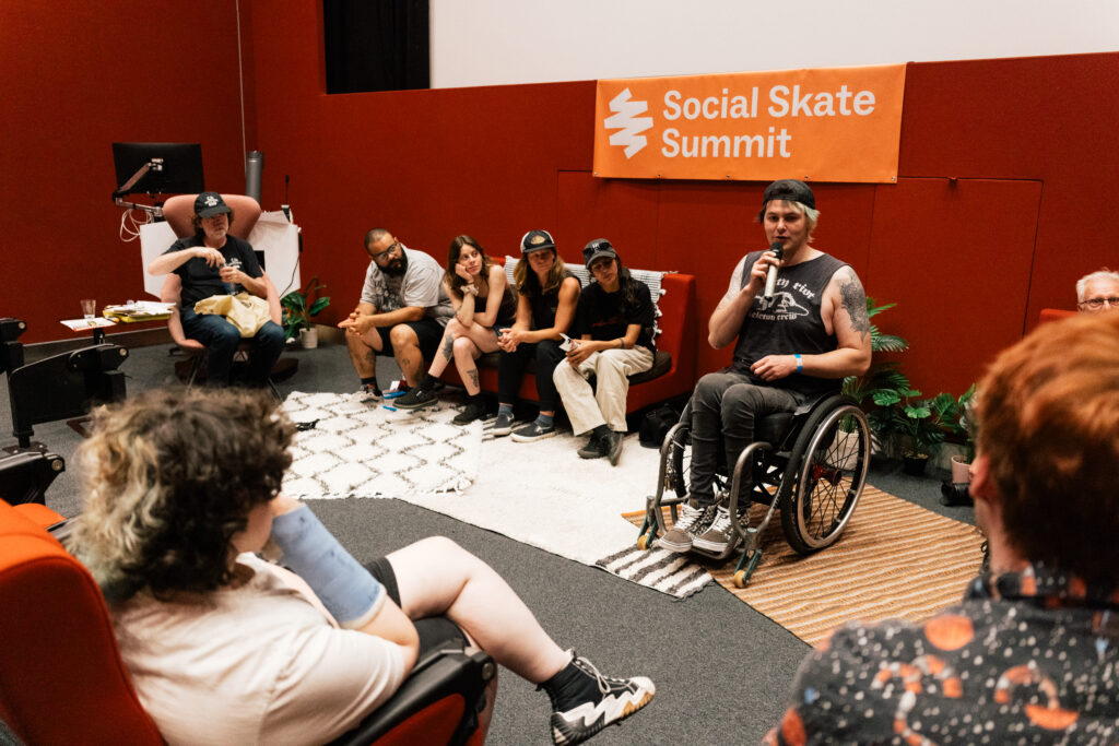 David sitzt im Rollstuhl auf einer Bühne beim Social Skate Summit in London und spricht in ein Mikrofon. Hinter ihm sitzen weitere Teilnehmende einer Diskussionsrunde auf Sofas und Sesseln. Im Vordergrund hören mehrere Personen aufmerksam zu. Über der Bühne hängt ein orangefarbenes Banner mit der Aufschrift „Social Skate Summit“.