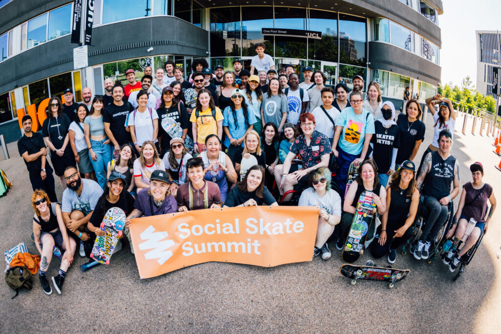 Rund 60 Teilnehmende des Social Skate Summit versammeln sich vor einem Gebäude der UCL in London für ein gemeinsames Gruppenfoto. Die Gruppe besteht aus Menschen unterschiedlicher Herkunft, Geschlechter und Altersgruppen. Einige halten Skateboards, mehrere Personen nutzen Rollstühle. Vor der Gruppe wird ein großes orangefarbenes Banner mit der Aufschrift „Social Skate Summit“ gehalten.