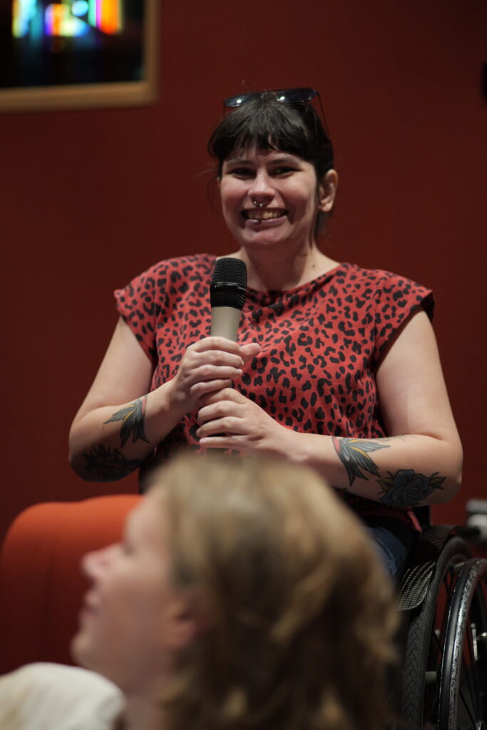 LIsa als Teilnehmerin sitzt im Rollstuhl und spricht lächelnd in ein Mikrofon. Sie trägt ein rot-schwarz gemustertes Oberteil und hat Tätowierungen an den Armen. Im Hintergrund ist die rote Wand des Veranstaltungsraums zu sehen, im Vordergrund verschwommen weitere Zuhörende.