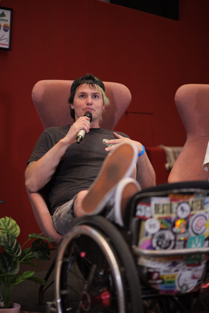David sitzt im Rollstuhl als Teil einer Podiumsdiskussion beim Social Skate Summit. Gemeinsam mit weiteren Teilnehmenden spricht er über Themen rund um Skateboarding, Inklusion und Community-Arbeit. Die Gesprächsrunde findet in einem kleinen Veranstaltungsraum mit Publikum statt.