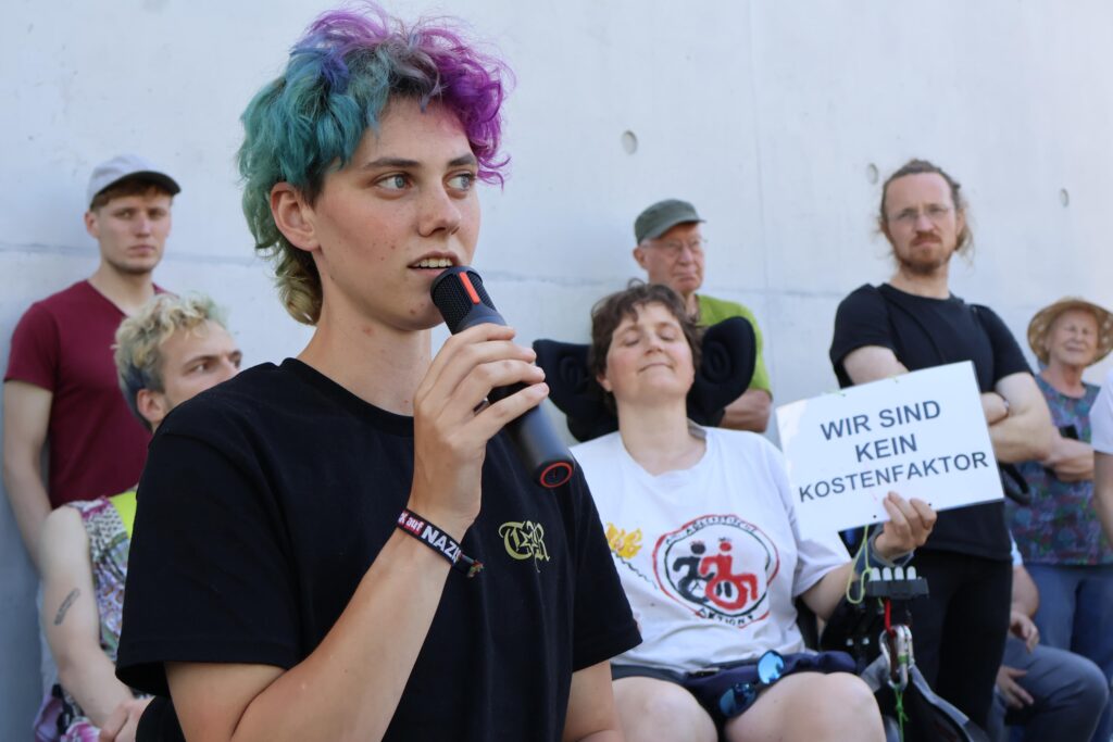 Eine Person mit türkis-lila Haaren spricht in ein Mikrofon. Hinter ihr hält eine Person im Rollstuhl ein Schild mit der Aufschrift „Wir sind kein Kostenfaktor“.