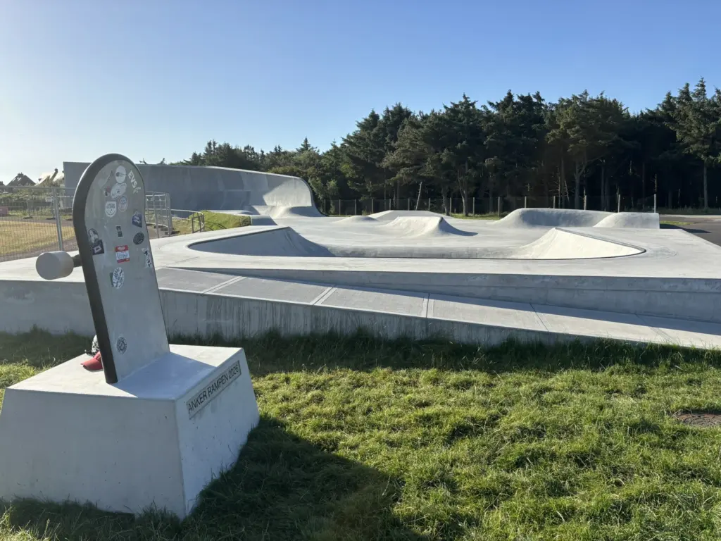 Zu sehen ist ein Skatepark im Hintergrund. Vorn am Skatepark ist eine Rampe für Rollstuhlfahrende Menschen. Im Vordergrund etwas Wiese auf dem eine Skateboard Skulptur steht.