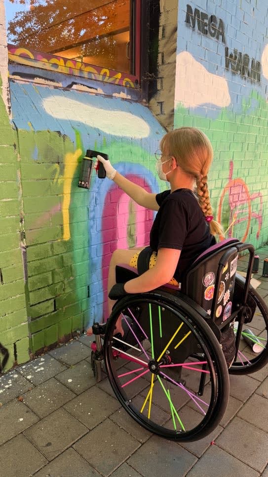 Ein Mädchen im Rollstuhl an einer Wand mit Graffiti Dose malt damit einen Regenbogen.
