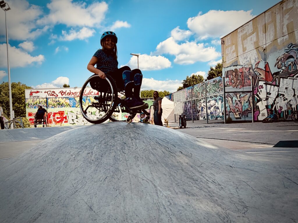 Ein Mädchen im Rollstuhl fährt über einen Beton Wobbel im Skatepark.