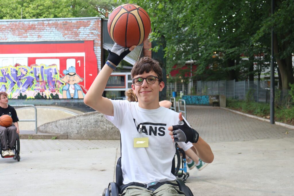 Ein Jugendlicher im Rollstuhl hält mit einer Hand einen Basketball über den Kopf. Im HIntergrund weitere Jugendliche mit Rollstuhl und Basketball.