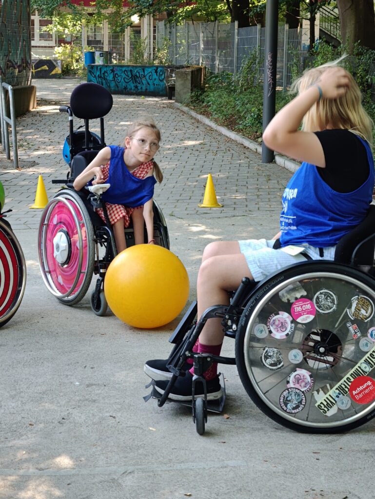 Eine Jugendliche im Rollstuhl steuert mit ihrer Hand am Joystick den Rollstuhl und die andere Hand führt einen gelben Gymnastikball. Davor steht eine weitere Jugendliche im Rollstuhl.