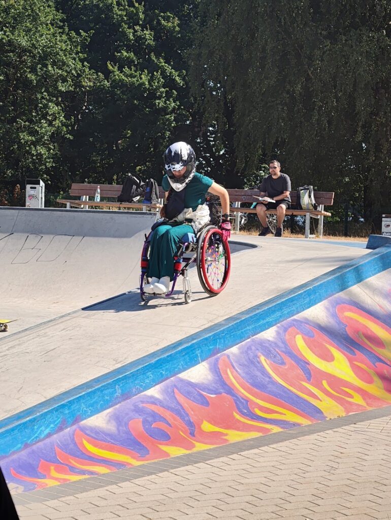 Eine Rollstuhlfahrerin fährt eine Rampe im Skatepark herunter. Die Rampe ist mit Graffiti Flammen verziert.
