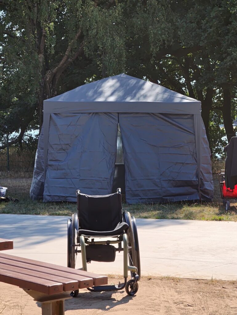 Ein Pavillon für das Camping Klo neben dem Skatepark. Im Vordergrund steht ein Rollstuhl im Skatepark.