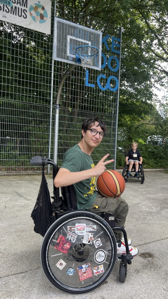 Jugendlicher im Rollstuhl hält einen Basketball und macht eine Peace Geste und steht mit seinem Rollstuhl unter einem Basketball Korb.